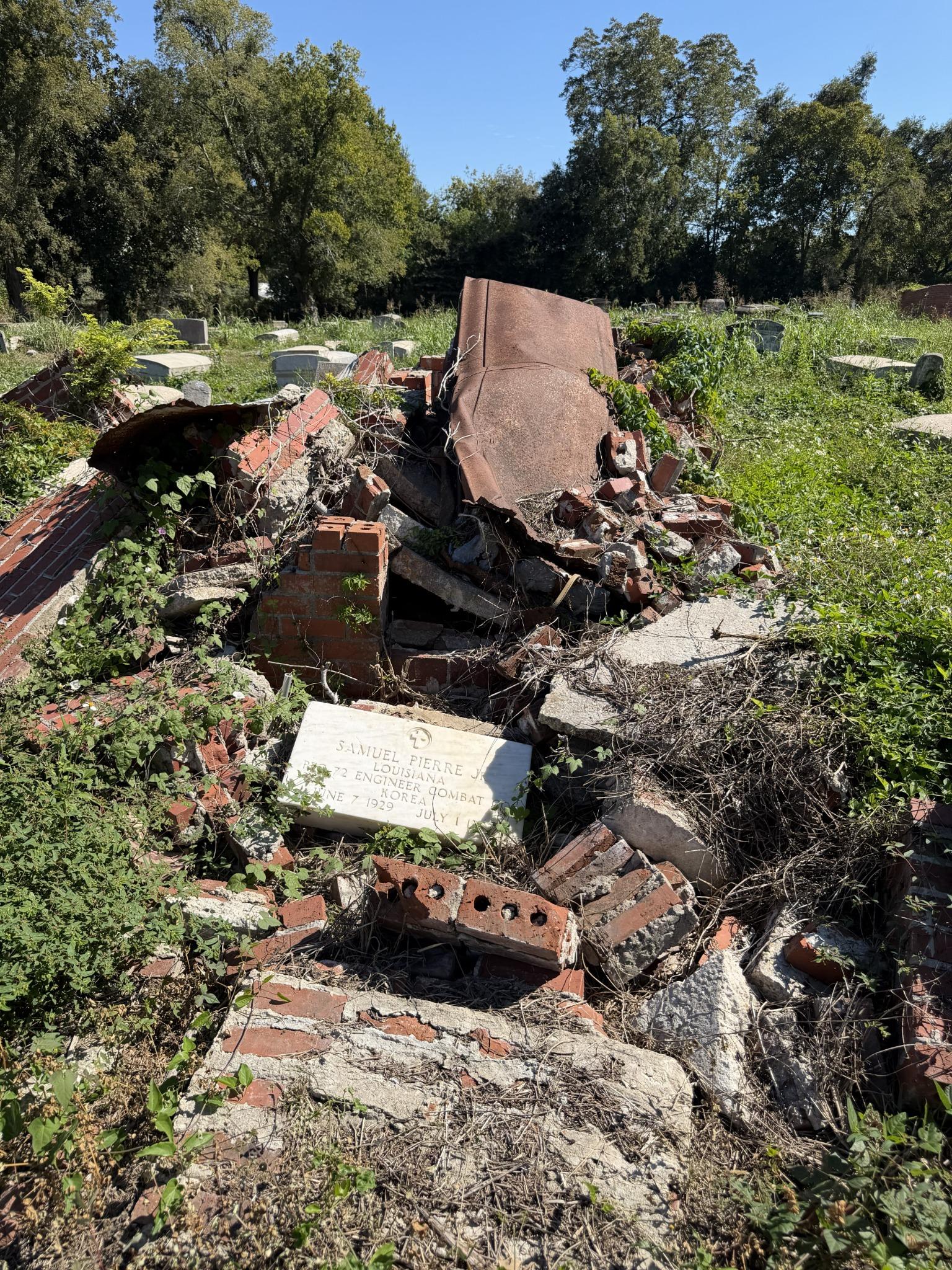 Pierre tombale d'un vétéran au milieu de briques brisées, de débris de béton et de végétation envahissante dans un cimetière, avec d'autres pierres tombales en arrière-plan.